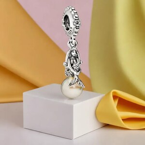 Puede incluir: Un charm de plata con un dise&ntilde;o de sirena y una perla. El charm presenta una figura detallada de sirena con una cola fluida, sosteniendo un ramo. El charm est&aacute; suspendido de una parte superior decorativa con detalles transparentes.