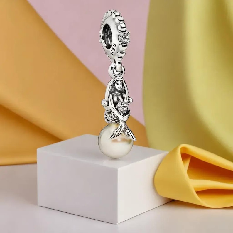 Puede incluir: Un charm de plata con un dise&ntilde;o de sirena y una perla. El charm presenta una figura detallada de sirena con una cola fluida, sosteniendo un ramo. El charm est&aacute; suspendido de una parte superior decorativa con detalles transparentes.