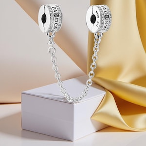 Può includere: Una catena di sicurezza per charm Pandora in argento. Due charm rotondi, ciascuno con il logo Pandora e incrostato di piccole pietre trasparenti, sono collegati da una catena. La catena è esposta su una scatola bianca con sfondo in raso dorato.