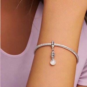 Puede incluir: Una pulsera plateada con una banda texturizada y un dije colgante. El dije presenta un peque&ntilde;o dise&ntilde;o adornado con una piedra blanca y redonda en la parte inferior. La pulsera se usa en un brazo sobre un fondo morado claro.