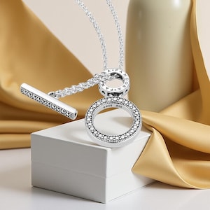 May include: A silver necklace featuring a round pendant and a T-bar clasp. The round pendant is adorned with small, clear stones. The necklace is presented on a white box with a gold ribbon in the background.