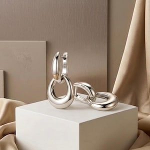Op de afbeelding: Een paar gepolijste zilveren oorringen. Elke oorring heeft een grote, druppelvormige hanger die is verbonden met een kleinere, cirkelvormige ring. De oorbellen worden gepresenteerd op een witte kubus tegen een neutrale achtergrond.