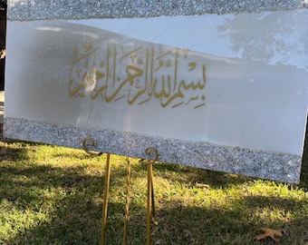 May include: A rectangular, white canvas with gold Arabic calligraphy, framed with a sparkling silver border. The artwork is displayed on a gold easel outdoors, set against a backdrop of green grass and trees.