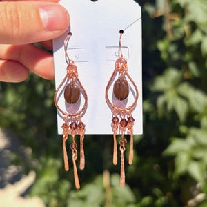 May include: A pair of copper-toned dangle earrings featuring a teardrop-shaped wire frame. Each earring contains a single dark brown coffee bean at the centre, accented with small, faceted brown crystal beads and thin, flat copper-toned metal dangles.