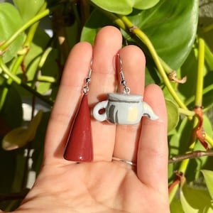 May include: A pair of handmade polymer clay earrings. One earring is a burgundy cone shape and the other is a gray and white teapot.