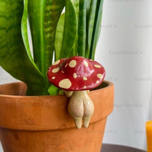 May include: A whimsical red mushroom plant pot decoration with white spots, perched on the edge of a terracotta pot. The mushroom has a beige stem and is next to a green plant.