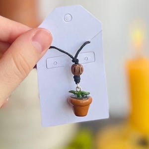 May include: A handmade necklace featuring a miniature potted plant pendant. The pendant is a small, brown pot with green leaves. The necklace has a black cord and a wooden bead accent.