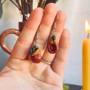 Puede incluir: Pendientes hechos a mano con diseños de cactus y manzana en miniatura. El pendiente de cactus tiene un cactus verde en una maceta marrón con hojas naranjas. El pendiente de manzana es rojo con un lazo naranja. Ambos pendientes tienen ganchos plateados.
