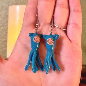 May include: A pair of blue pig-shaped earrings with brown button noses. The earrings are made of polymer clay and have silver hooks.