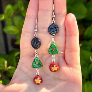 May include: A pair of handmade earrings featuring a unique design. Each earring has a silver hook, a black button, a green triangle, and a red sphere with a yellow star. The earrings are suspended by silver chains.