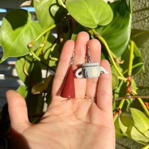 May include: A pair of handmade polymer clay earrings. One earring is a red cone shape and the other is a gray teapot with a yellow flower design.