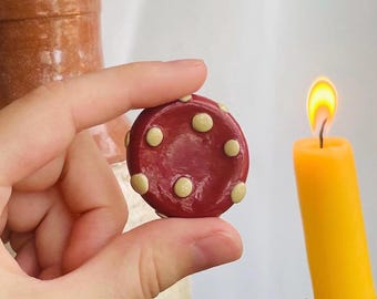 May include: A small, round, red mushroom-shaped object with cream-colored spots is held between fingers. A yellow candle with a lit flame is in the background. The mushroom object appears to be handmade.