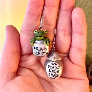 Puede incluir: Un par de pendientes de plata con una rana verde en un frasco que dice "Frog's Breath" y un frasco de plata que dice "Deadly Night Shade".
