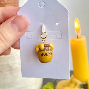 May include: A miniature yellow honey pot charm with the word "HUNNY" in brown lettering. The charm is attached to a white tag with a gold-colored ring. A lit yellow candle is in the background.
