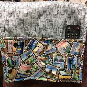 Pode incluir: Um encosto de cadeira de tecido cinza e branco tecido com um bolso de tecido castanho. O bolso tem uma estampa colorida de selos de estado com os nomes de estados como Carolina do Sul, Idaho, Texas e Oklahoma. Um controle remoto preto está no bolso.