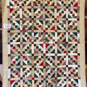 Könnte beinhalten: Handgefertigte Patchwork-Quilt mit einem Muster aus Quadraten, Dreiecken und Rechtecken in verschiedenen Farben wie Rot, Grün, Blau und Braun. Der Quilt hat einen beigen Rand mit roter Zierleiste. Ein gemütliches Wohnaccessoire.