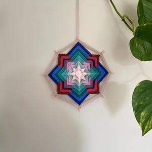 Op de afbeelding: Een zeshoekige wandhanger gemaakt van garen gewikkeld rond houten stokjes. Het ontwerp bevat concentrische geometrische lagen in tinten lichtroze, magenta, taling, koningsblauw en donkerbruin, wat een stervormig patroon creëert.