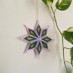 May include: A handmade star-shaped wall hanging. The star features a geometric design with alternating sections of purple, green, and yellow yarn. The star is suspended by a pink string against a white wall, with green and white leafy vines in the background.