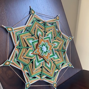 May include: A multi-layered yarn mandala featuring a geometric star pattern in shades of forest green, burnt orange, and white. The intricate design is woven onto a wooden frame with six points, creating a symmetrical, layered, and textured appearance.