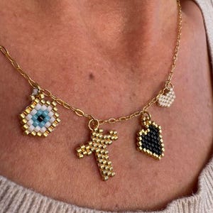 Puede incluir: Collar de cadena dorada con múltiples colgantes de cuentas. Los colgantes incluyen un ojo, una cruz, un corazón y un pequeño colgante en forma de corazón. El colgante del ojo tiene detalles azules y blancos. Los colgantes de cruz y corazón son dorados.