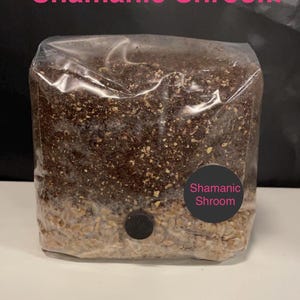 May include: A clear plastic bag filled with brown and white grains. A black sticker with the text "Shamanic Shroom" is on the bag.