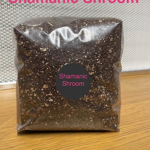 May include: A transparent plastic bag containing dark brown material, possibly compost or a growing medium. The text "Shamanic Shroom" is displayed in pink at the top and on a circular label.