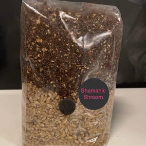 May include: A clear plastic bag containing a brown substrate and a layer of wheat berries. A black sticker with the text "Shamanic Shroom" is attached to the bag.
