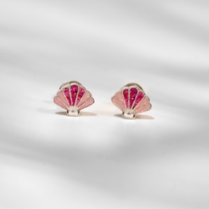 May include: A pair of stud earrings shaped like seashells. Each earring features a silver-toned metal outline with alternating sections of light pink enamel and vibrant, glittery magenta accents, creating a distinct fan-like shell design.