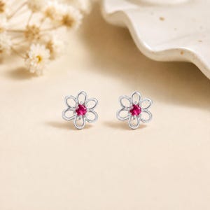 May include: A pair of silver stud earrings shaped like six-petaled flowers. Each earring features a small, round, vibrant pink gemstone set in the center of the silver floral frame, creating a delicate and symmetrical appearance for the jewelry.