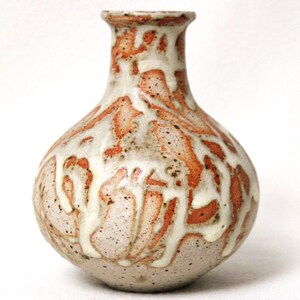 May include: A small, white ceramic vase with a brown and orange glaze. The glaze has a drip effect, creating a unique pattern.
