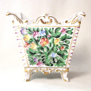 Puede incluir: Jardinera de porcelana blanca con diseño floral. La jardinera tiene forma cuadrada con adornos dorados, asas y patas. La parte delantera está decorada con un colorido arreglo floral de flores rojas, amarillas, rosas y azules.