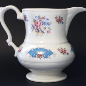 Può includere: Un'anfora di ceramica bianca con un motivo floreale. L'anfora ha un motivo floreale blu e bianco intorno alla base e un motivo floreale più grande sul corpo. L'anfora ha un manico e un beccuccio.