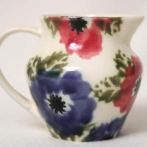 May include: A white ceramic jug with a floral design. The jug has a handle and is decorated with blue, red and green flowers.