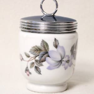 Op de afbeelding: Een wit porseleinen eierdopje met een zilveren deksel en een bloemmotief met paarse en roze bloemen. Het eierdopje heeft een zilveren handvat.