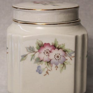 May include: White ceramic jar with a gold trim and floral design. The jar has a lid and is decorated with pink, purple, and blue flowers.