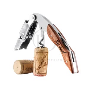 May include: A stainless steel and wood wine opener with a corkscrew, lever, and bottle opener. The wooden handle is engraved with the name "Michael". Two corks are also present, one with the text "GOLDWAHA" and the other with "FRANCE".