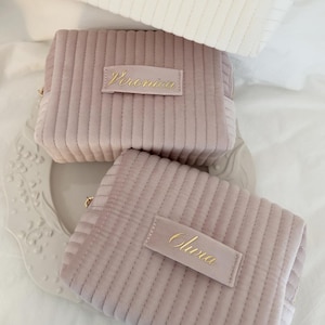May include: Three corduroy cosmetic bags in soft colors. The top bag is white with "Victoria" in gold script. The middle bag is mauve with "Veronica" in gold script. The bottom bag is mauve with "Olivia" in gold script.