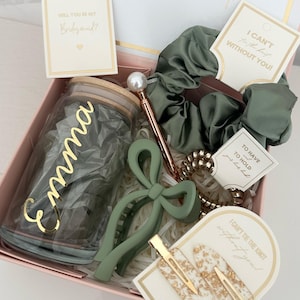 May include: A pink gift box filled with green and gold accessories. The box includes a glass jar with the name "Emma" written on it, a green hair clip, a green scrunchie, a gold hair clip, and several small gift tags with text that says "Will you be my bridesmaid?" and "I can't tie the knot without you!"