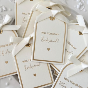 Op de afbeelding: Ivoor cadeaulabels met gouden rand en de tekst "Will You Be My Bridesmaid?" en "Will You Be My Matron of Honor?" zijn vastgebonden met ivoorkleurige satijnen linten. Elk label heeft een gouden oogje en een klein gouden hart.
