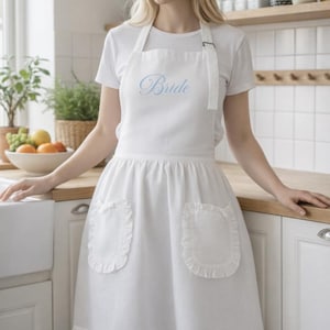 Peut inclure: Tablier blanc avec poches et ourlet à volants. Le mot "Bride" est brodé en script bleu clair sur la poitrine. Le tablier est porté sur un t-shirt blanc. Cadre de cuisine.