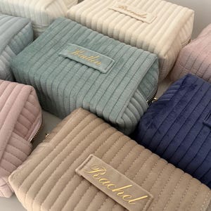 Puede incluir: Una colecci&oacute;n de neceseres de pana en varios colores, incluyendo rosa, azul, beige y verde. Cada bolsa tiene una etiqueta rectangular con un nombre bordado en escritura dorada. Las bolsas tienen cierre de cremallera y una textura acanalada.