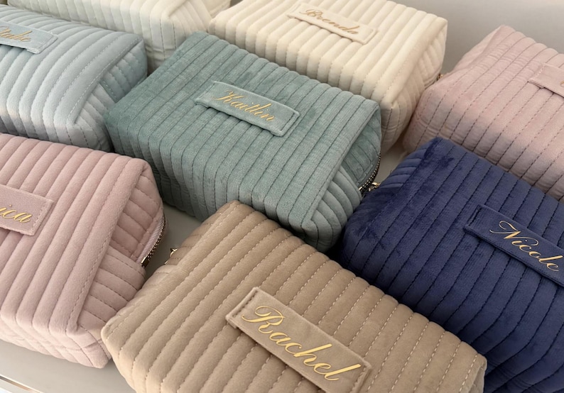 Puede incluir: Una colecci&oacute;n de neceseres de pana en varios colores, incluyendo rosa, azul, beige y verde. Cada bolsa tiene una etiqueta rectangular con un nombre bordado en escritura dorada. Las bolsas tienen cierre de cremallera y una textura acanalada.