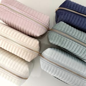 Puede incluir: Una colecci&oacute;n de estuches rectangulares acolchados en varios colores, incluyendo rosa, crema, azul y verde. Cada estuche tiene un cierre de cremallera dorado. Los estuches est&aacute;n colocados sobre una superficie blanca.