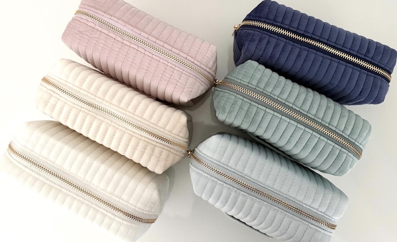 Puede incluir: Una colecci&oacute;n de estuches rectangulares acolchados en varios colores, incluyendo rosa, crema, azul y verde. Cada estuche tiene un cierre de cremallera dorado. Los estuches est&aacute;n colocados sobre una superficie blanca.