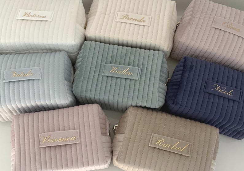 Puede incluir: Una colecci&oacute;n de neceseres de pana en varios colores pastel, incluyendo blanco, azul y rosa. Cada neceser presenta una etiqueta rectangular con un nombre bordado en dorado. Los neceseres tienen cierre de cremallera y una textura acanalada.