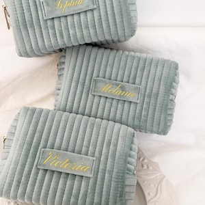 Puede incluir: Varias bolsas de cosm&eacute;ticos de pana verde azulado claro con cremalleras doradas y etiquetas de nombre personalizadas. Los nombres "Sophia", "Melanie" y "Victoria" est&aacute;n bordados en hilo dorado en las etiquetas rectangulares. Las bolsas est&aacute;n dispuestas sobre una superficie blanca.