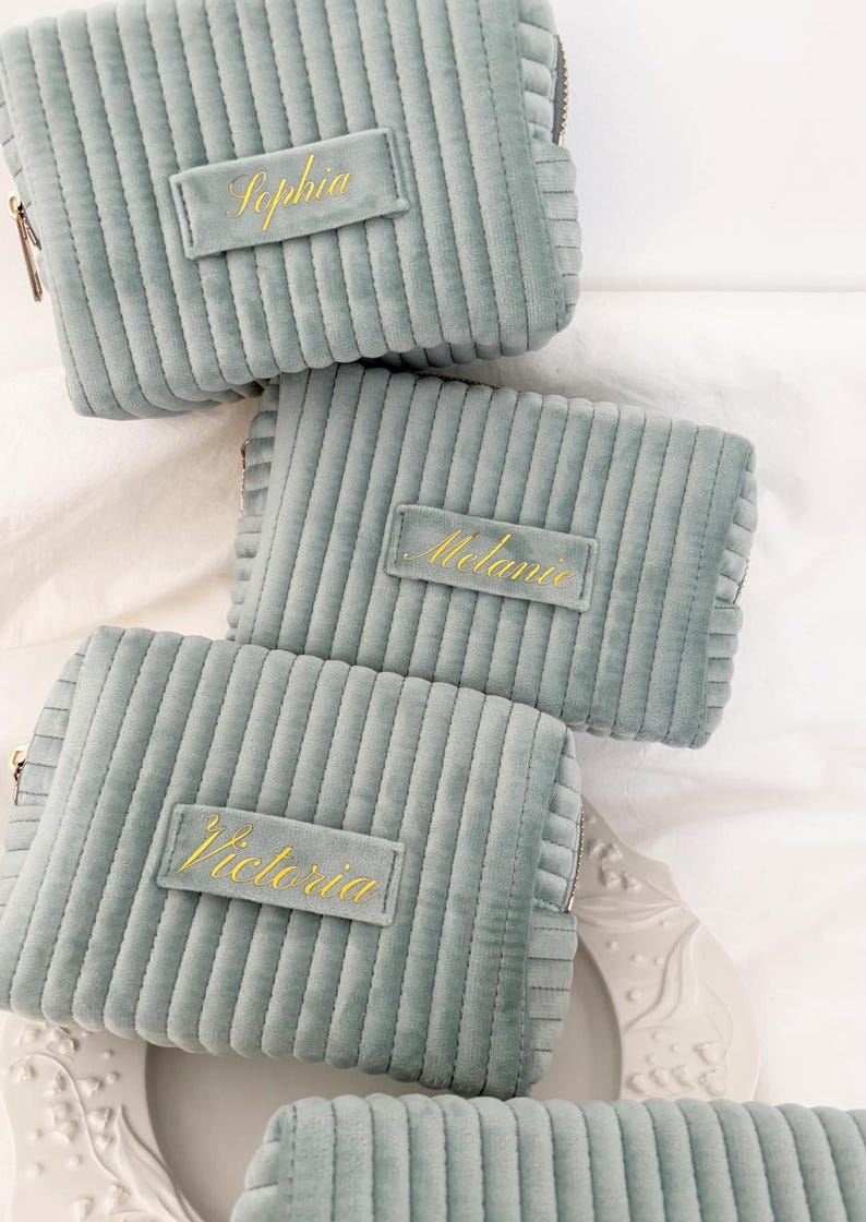 Puede incluir: Varias bolsas de cosm&eacute;ticos de pana verde azulado claro con cremalleras doradas y etiquetas de nombre personalizadas. Los nombres "Sophia", "Melanie" y "Victoria" est&aacute;n bordados en hilo dorado en las etiquetas rectangulares. Las bolsas est&aacute;n dispuestas sobre una superficie blanca.