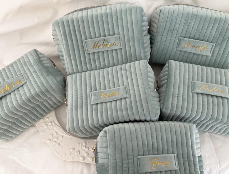 Puede incluir: Varias bolsas de cosm&eacute;ticos de pana azul claro, cada una con un nombre bordado en oro. Los nombres incluyen "Melanie", "Jennifer", "Victoria", "Sophie", "Heather" y "Tiffany". Las bolsas est&aacute;n dispuestas sobre una superficie blanca.