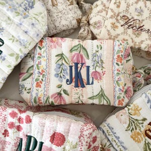 Può includere: Una collezione di pochette in tessuto trapuntato con vari motivi floreali nei toni del rosa, blu, giallo e verde. Ogni pochette è personalizzata con iniziali o nomi ricamati come JKL, Victoria e altri in filo scuro.