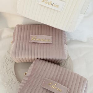 Puede incluir: Tres neceseres de pana en crema, malva y rosa empolvado. Cada bolsa tiene una etiqueta rectangular con un nombre en escritura dorada: "Victoria", "Veronica" y "Olivia". Las bolsas tienen cremallera y un dise&ntilde;o acolchado.